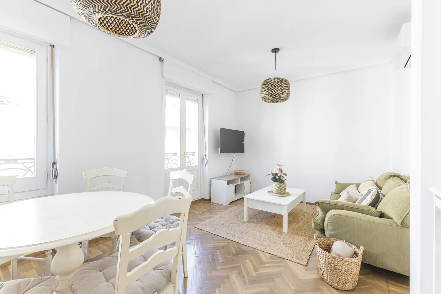Bright living room with white walls, herringbone wood floors, and woven light fixtures. A green sofa and white table add color.