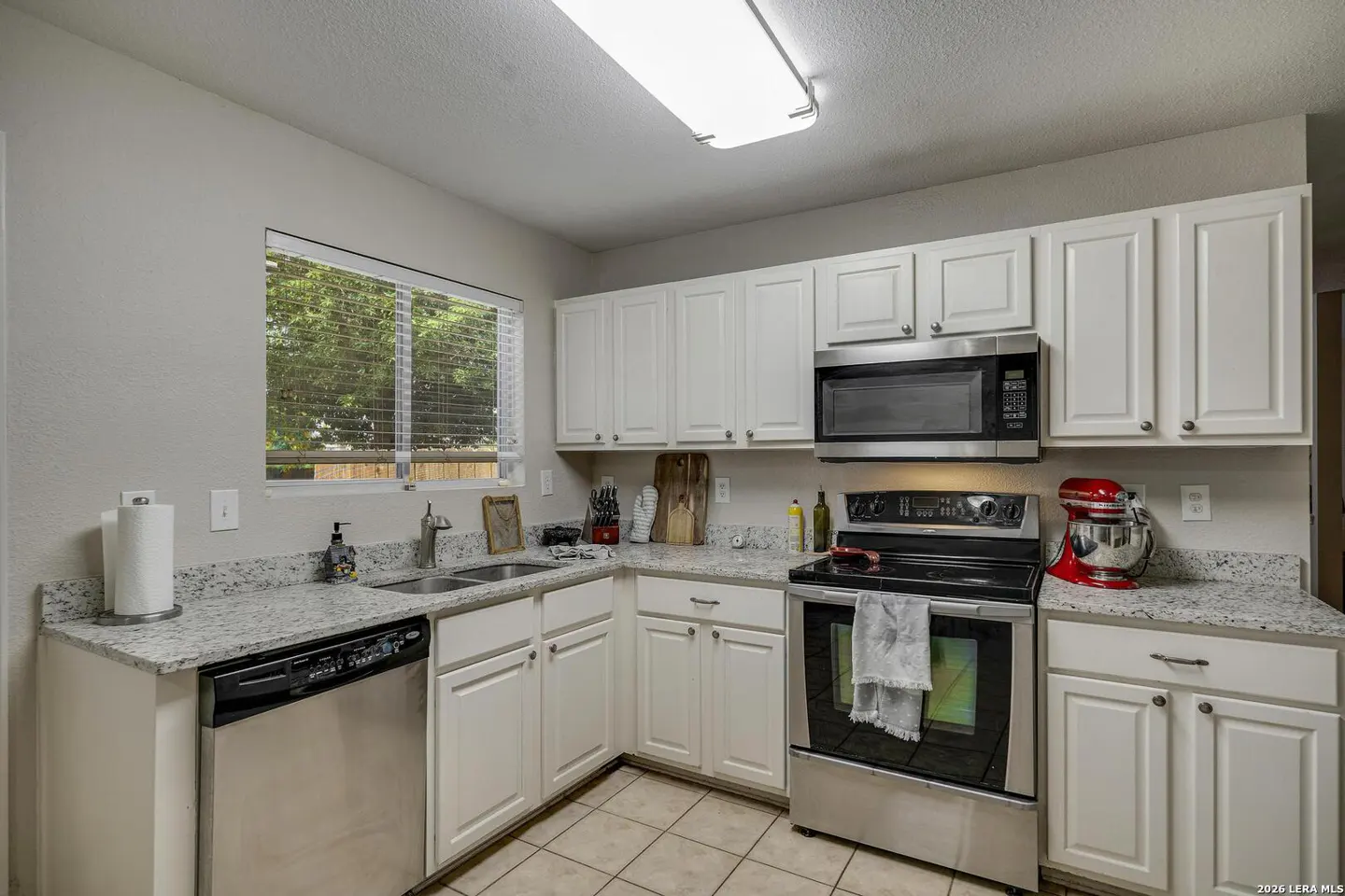 Bright kitchen with white cabinets, granite countertops, stainless steel appliances, and a window with blinds.