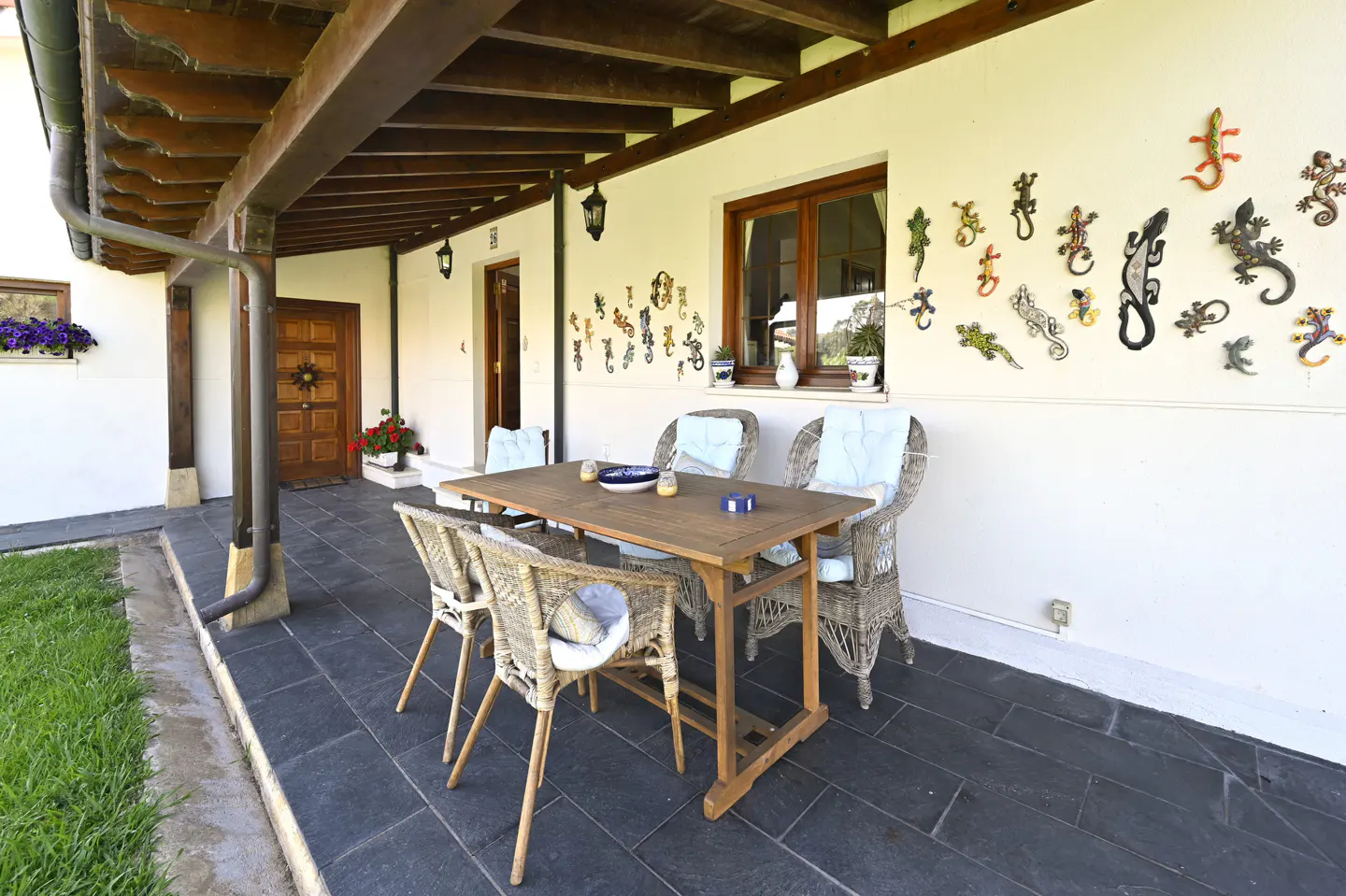 Covered patio with a wooden table and four wicker chairs with blue cushions. The white wall is decorated with metal lizards.