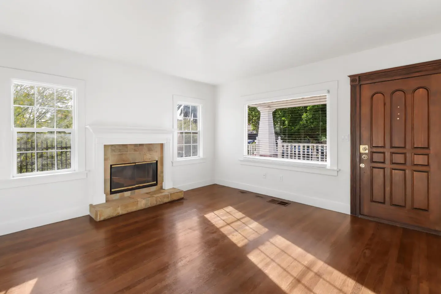 Bright living room with hardwood floors, white walls, and a stone fireplace. Windows offer outdoor views, and a wooden door adds warmth.
