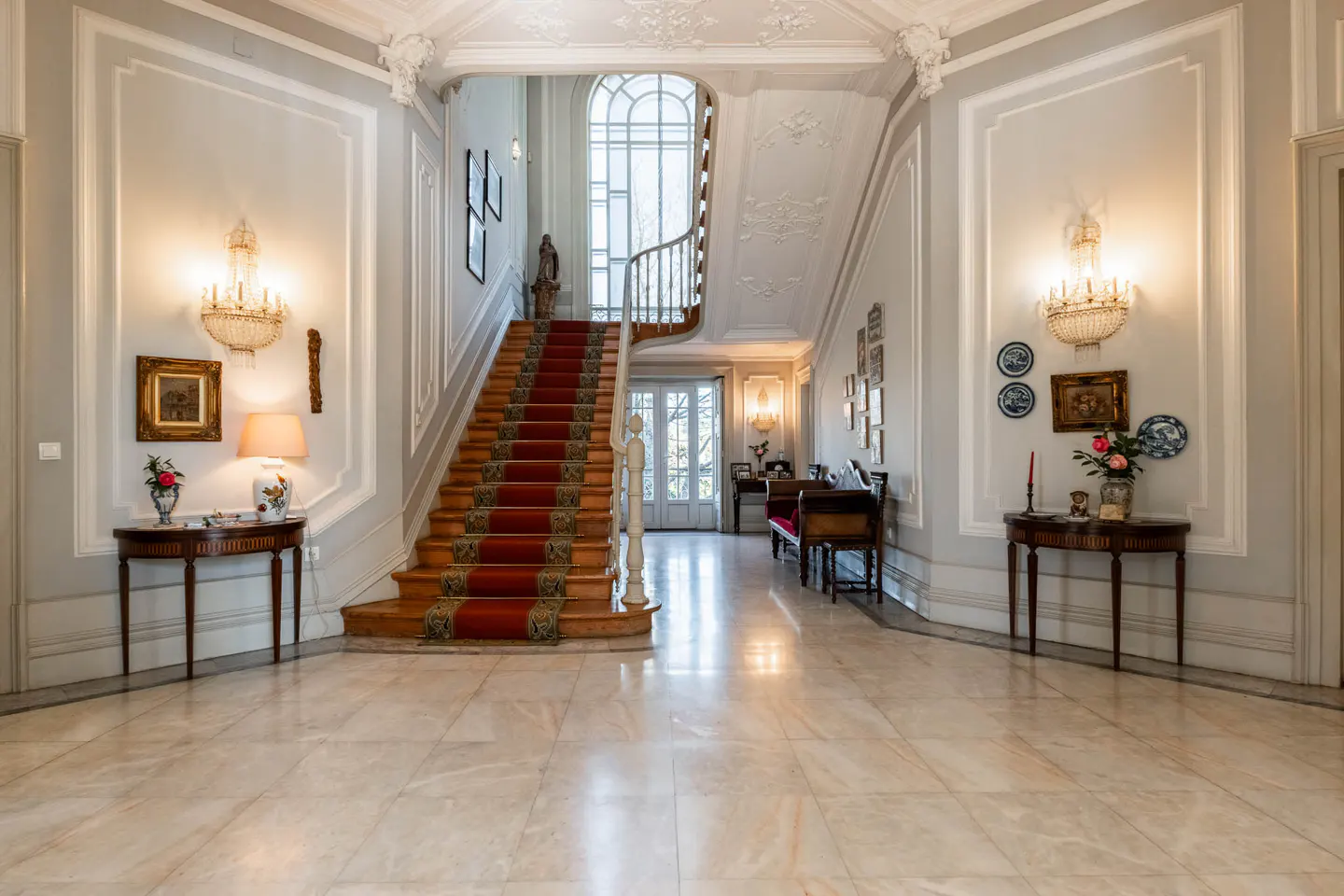 Elegant foyer with marble floors, light gray walls, and a staircase with a red runner. Chandeliers and artwork adorn the walls.