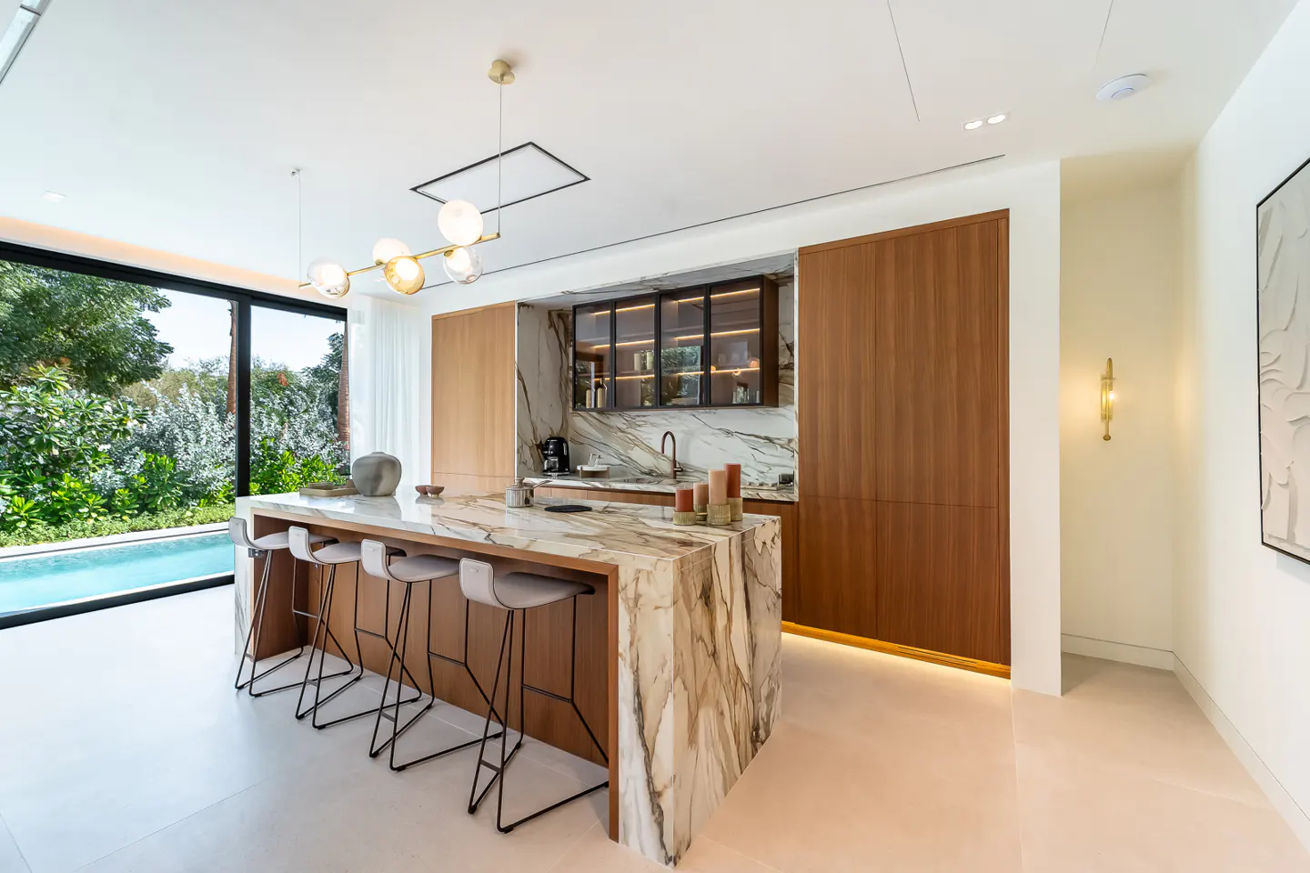 A modern kitchen with a marble island and bar stools, next to a pool visible through a large window.
