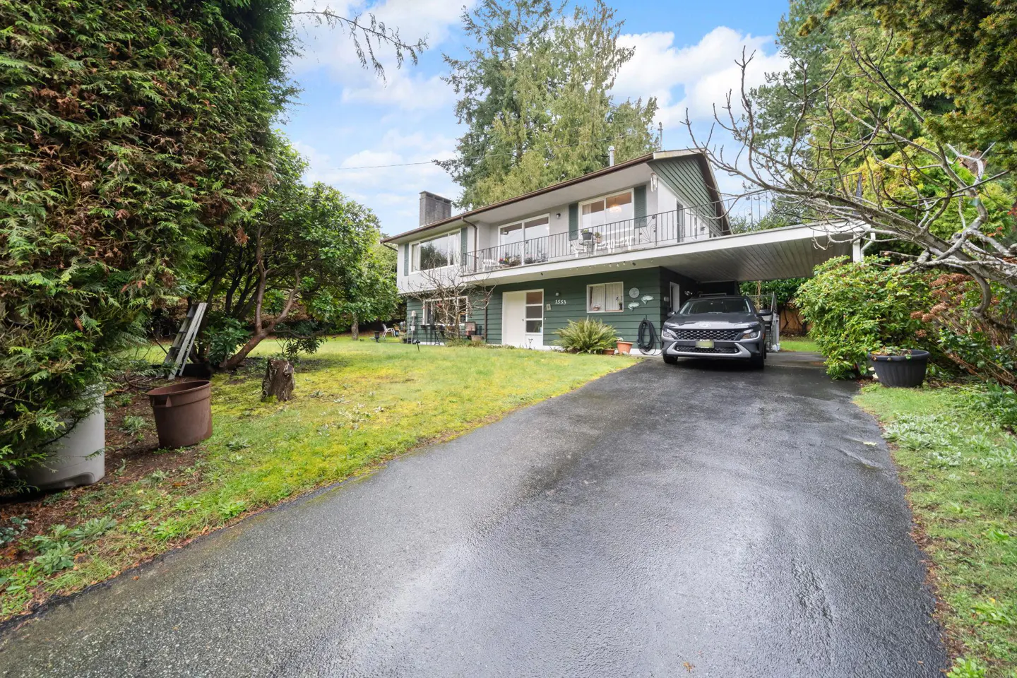 Two-story green house with a balcony and a car parked under a carport. A wet driveway leads to the house, surrounded by trees and grass.