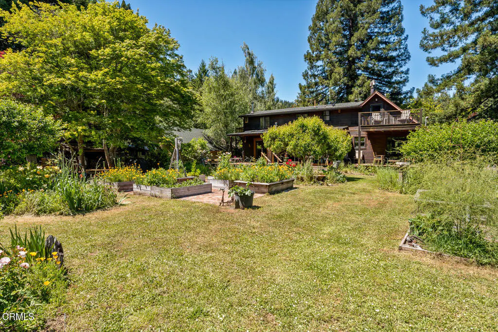 A lush green lawn with raised garden beds leads to a brown two-story house with a balcony, surrounded by trees under a blue sky.