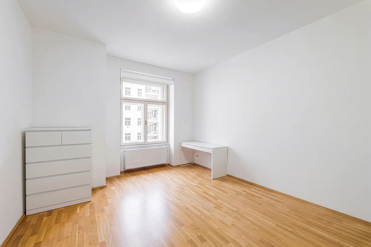 Bright, empty room with hardwood floors, white walls, a window, a white dresser, and a white desk.
