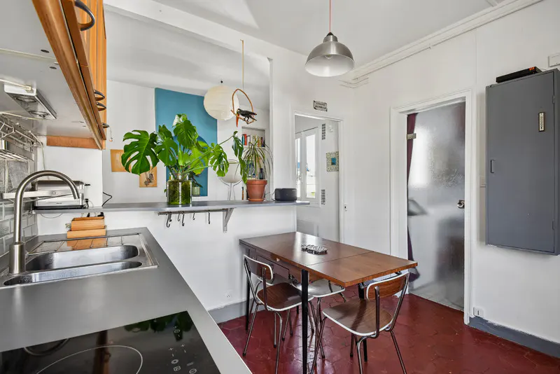 A bright kitchen with a stainless steel sink, dark countertop, and a wooden table with four chairs on a red floor.
