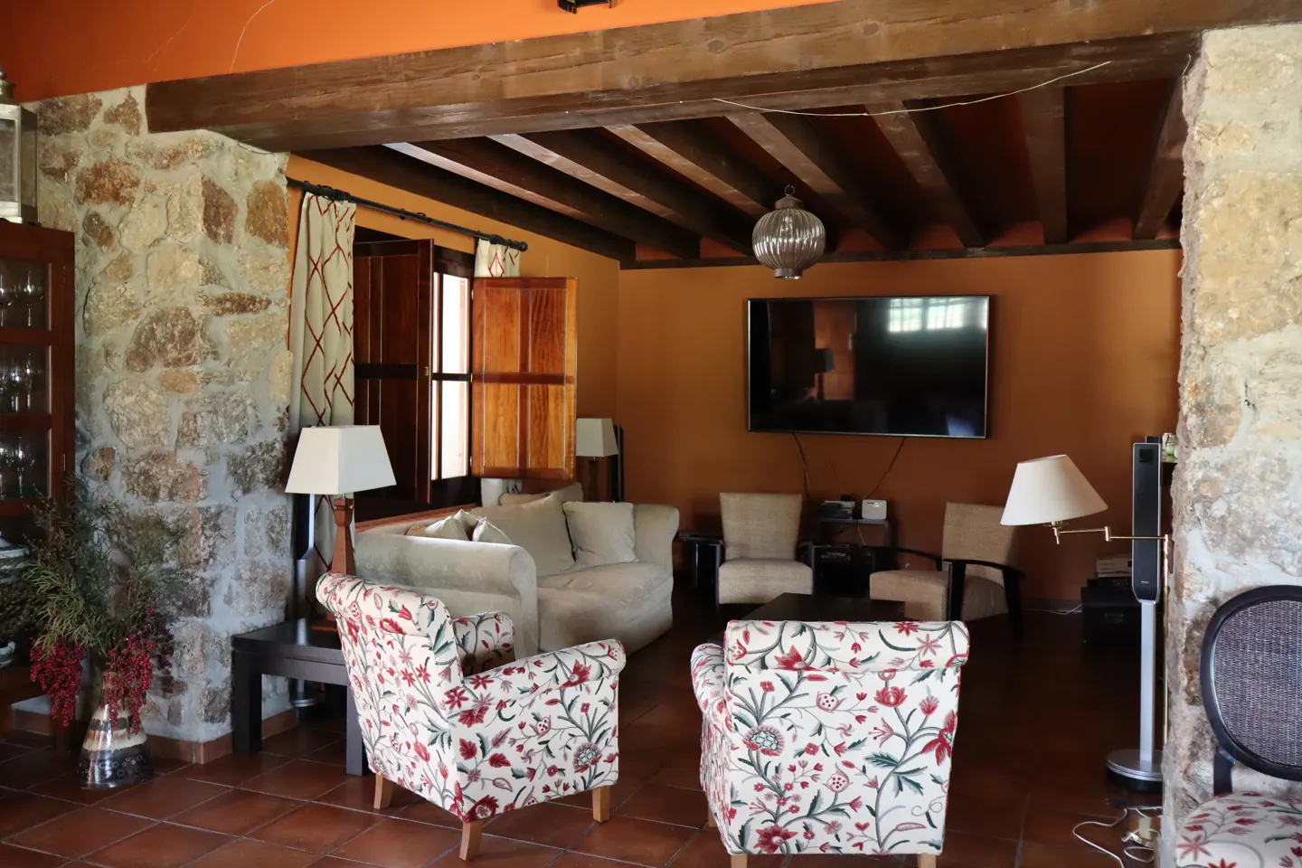 Living room with stone walls, wood beams, terracotta floors, floral chairs, and a cream sofa. A TV hangs on the orange wall.