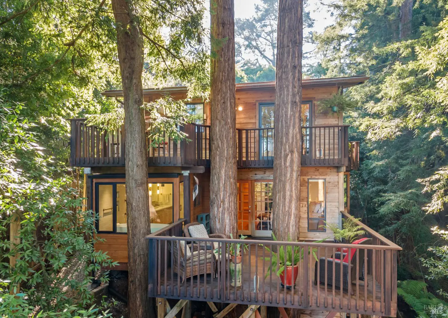 Two-story wood house with decks built around trees in a forest. The decks have railings and chairs.