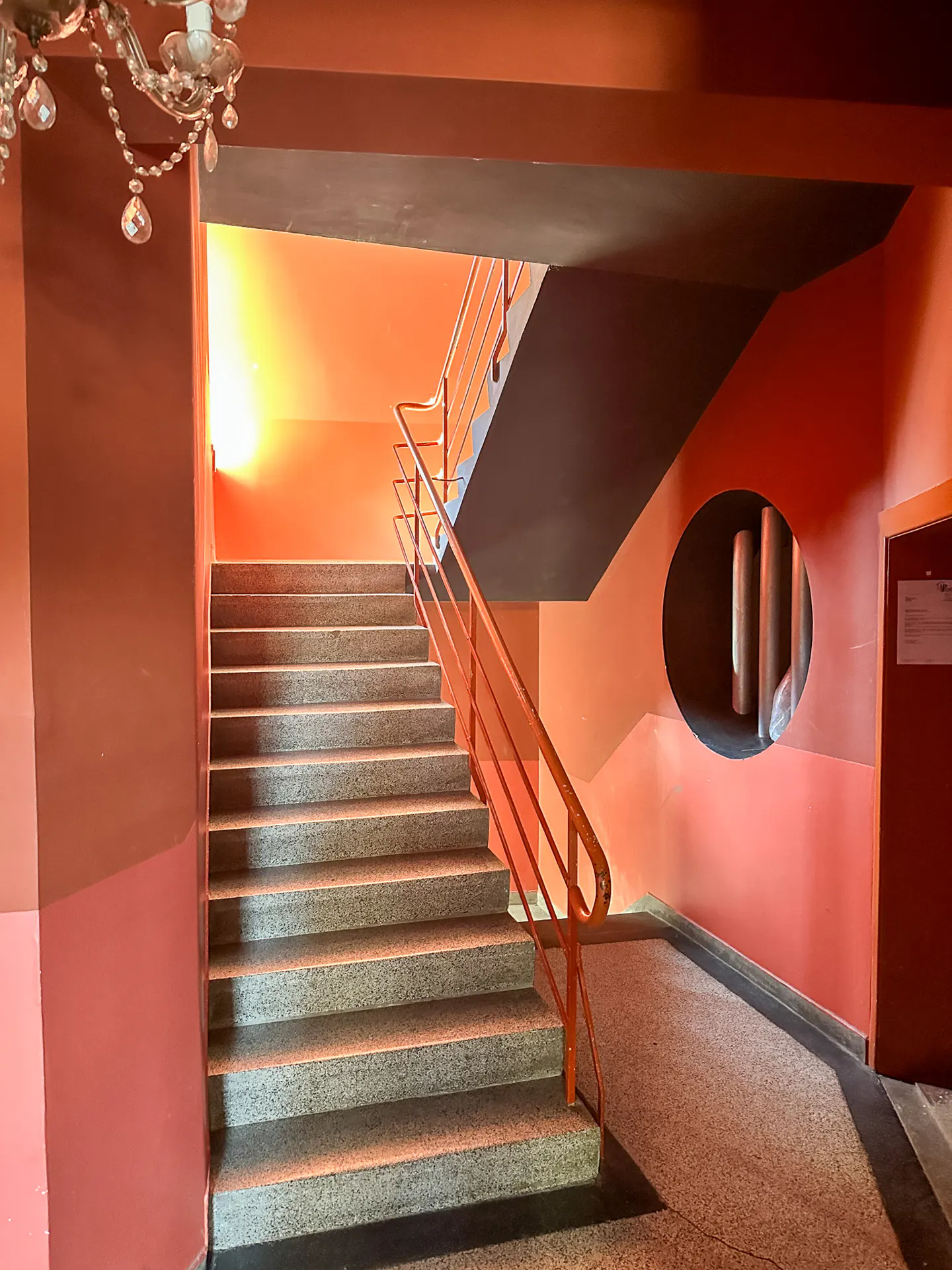 Staircase with stone steps and orange railings in a building with orange walls, a crystal chandelier, and a circular window.