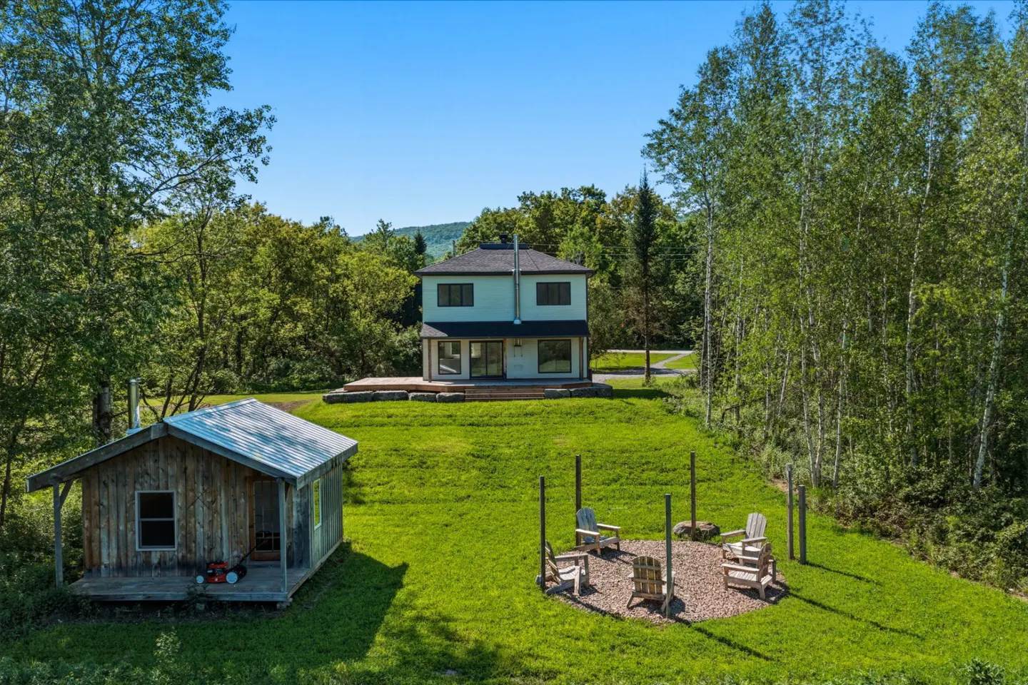 A two-story house, a rustic wooden shed, and a circular fire pit area with chairs sit on a lush green lawn near a forest.