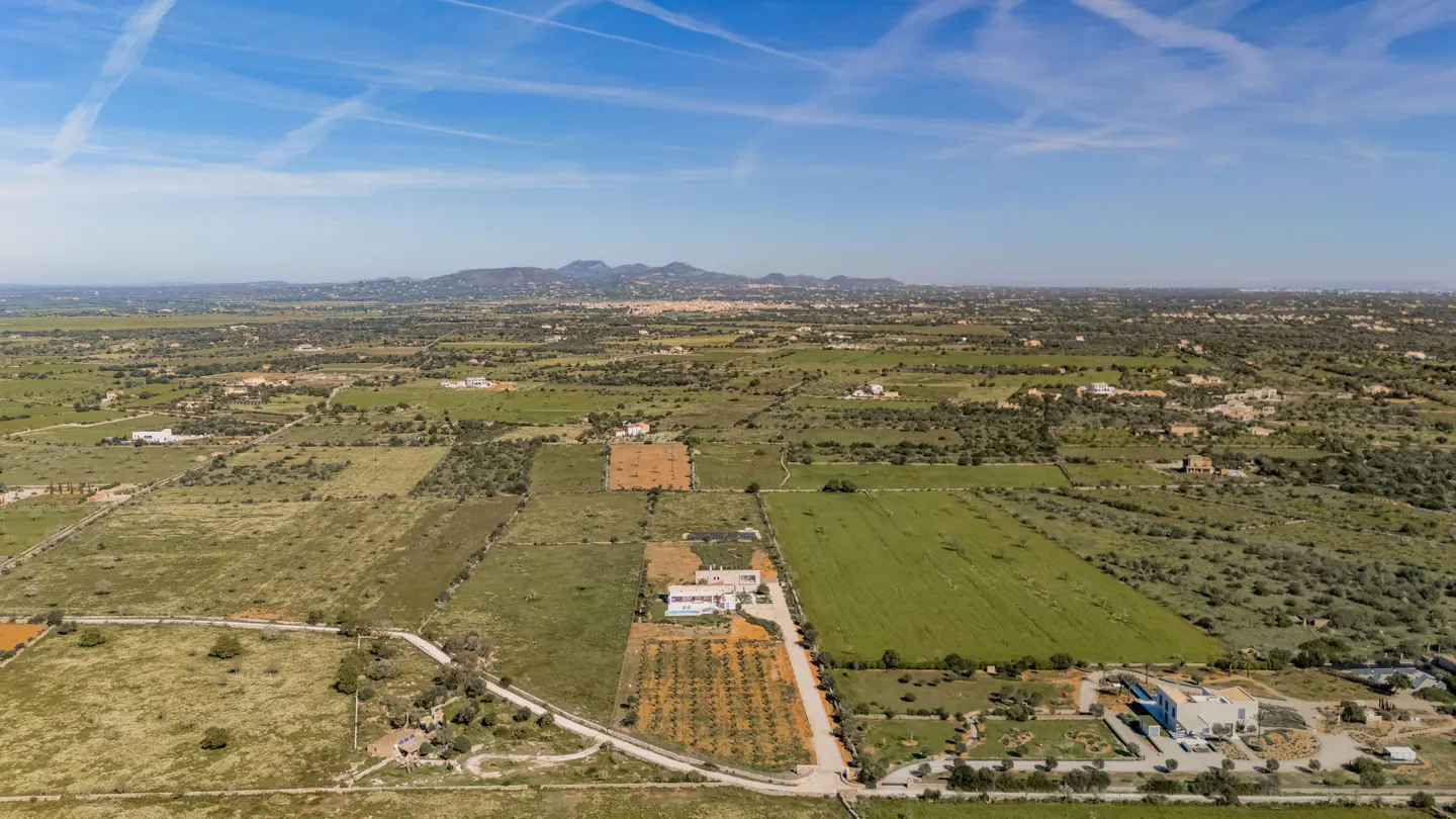 Aerial view of a rural landscape with green fields, scattered buildings, and distant mountains under a blue sky.