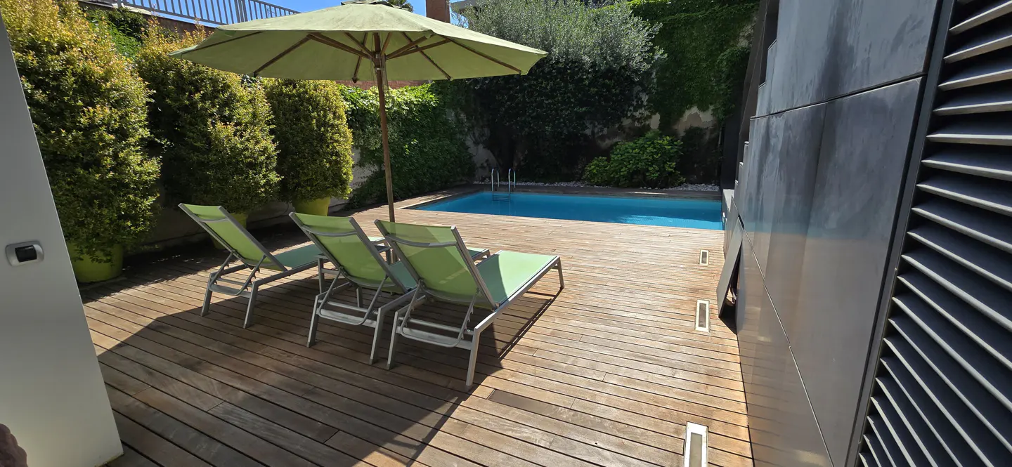 Outdoor pool area with wooden deck, three green lounge chairs, and a green umbrella. Lush greenery surrounds the pool.