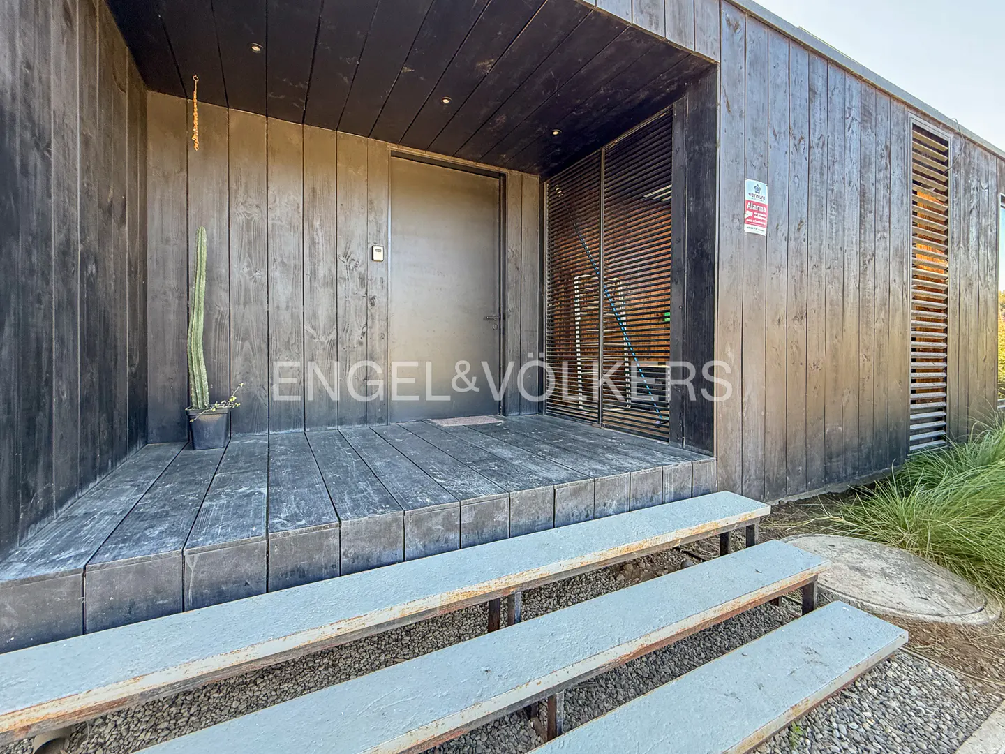 Exterior of a modern, dark wood home with a gray metal door and steps. A cactus sits on the porch.