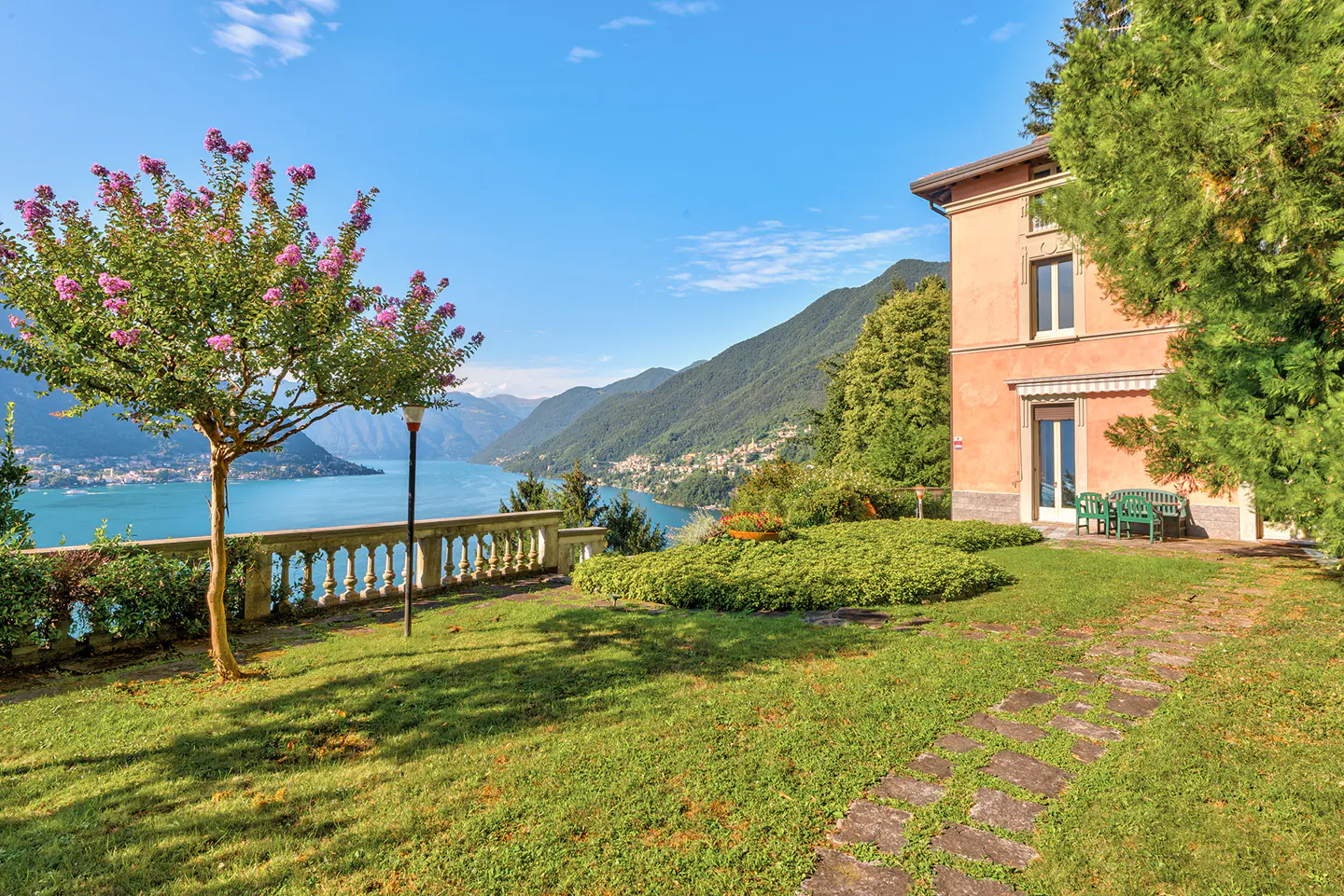 Scenic view of a peach-colored house with a green lawn overlooking a blue lake and mountains under a clear blue sky.