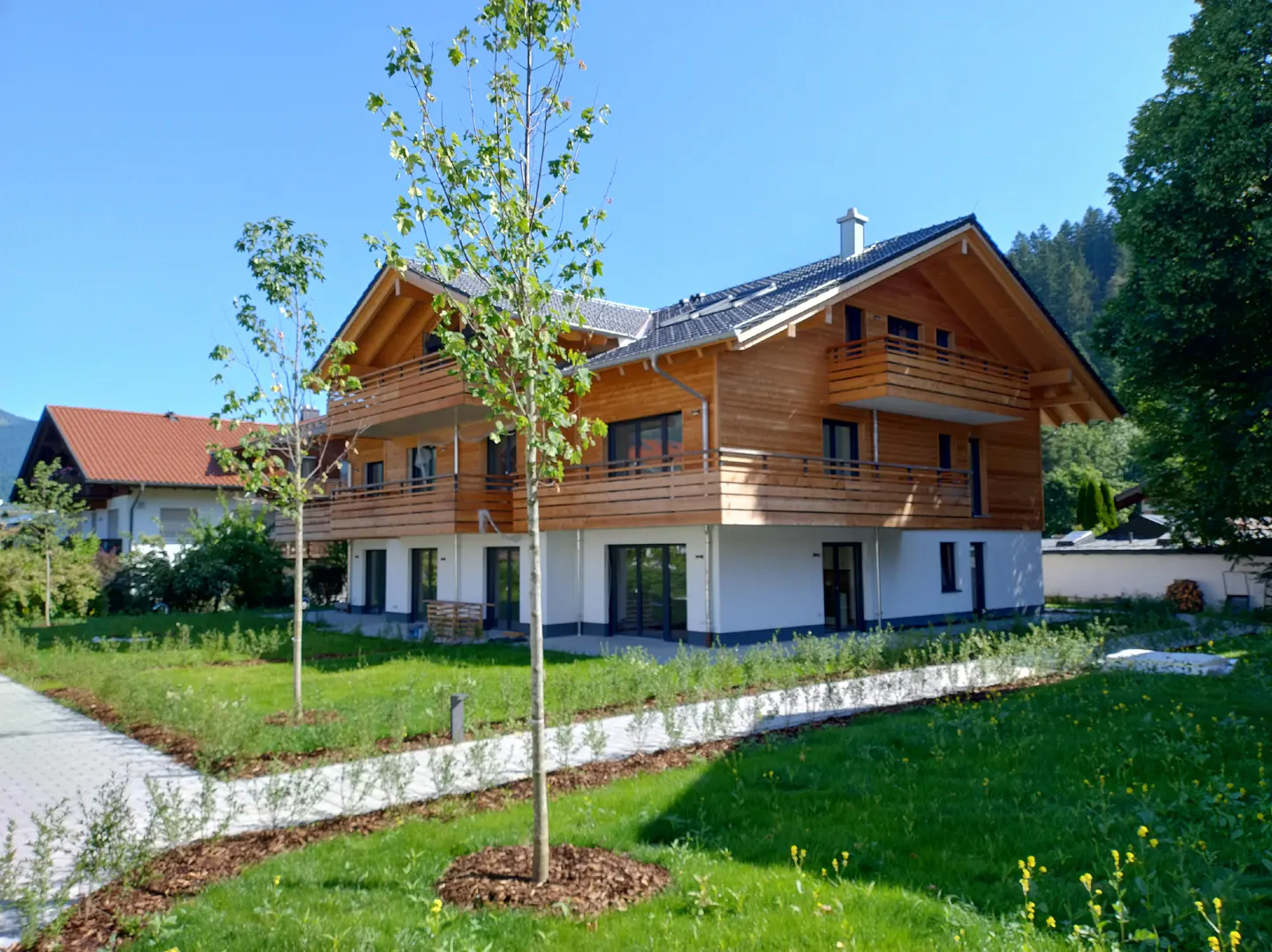 Two-story house with wood siding and balconies, set on a green lawn with young trees under a clear blue sky.