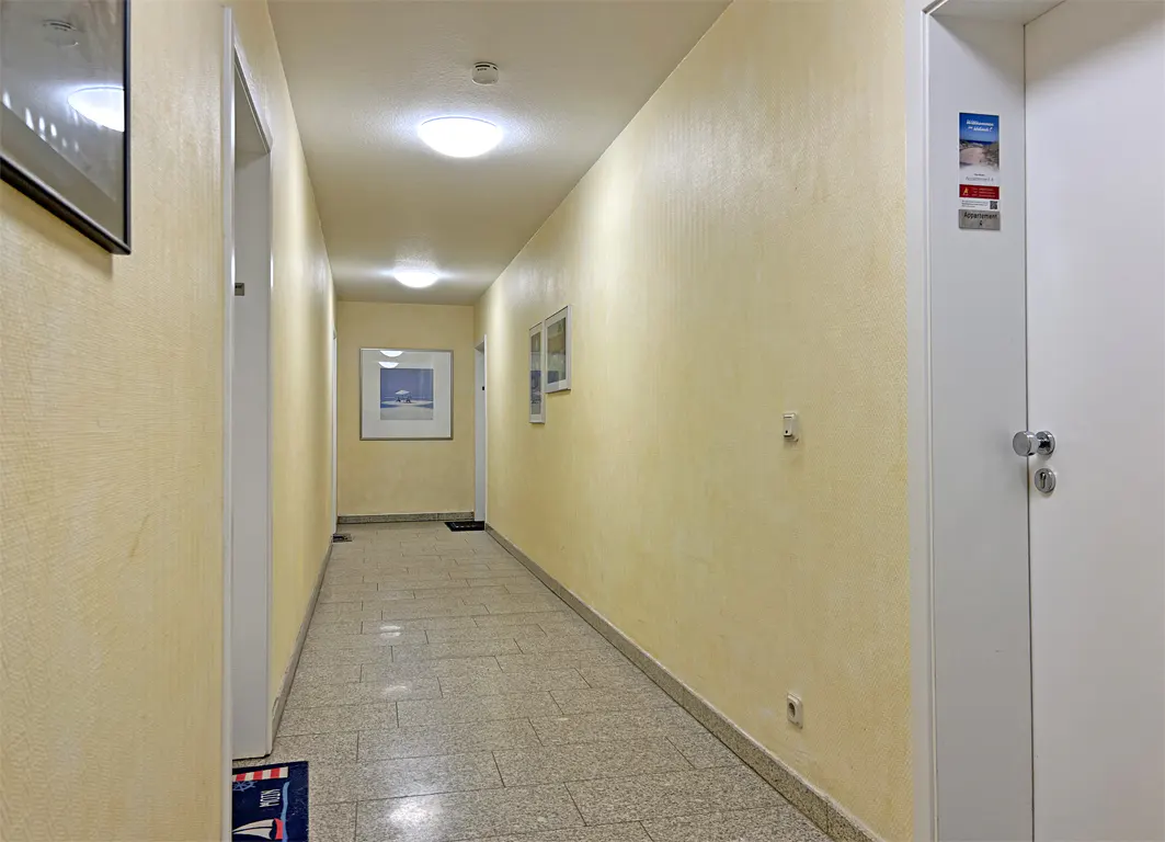 Hallway with yellow walls, tiled floor, and white doors. Pictures hang on the walls, and lights are on the ceiling.