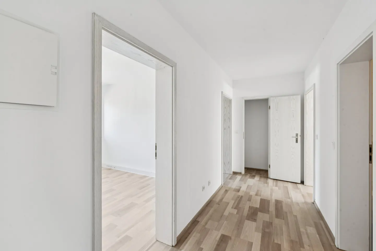 A bright hallway with white walls and light brown wood floors. Several open doorways lead to other rooms.