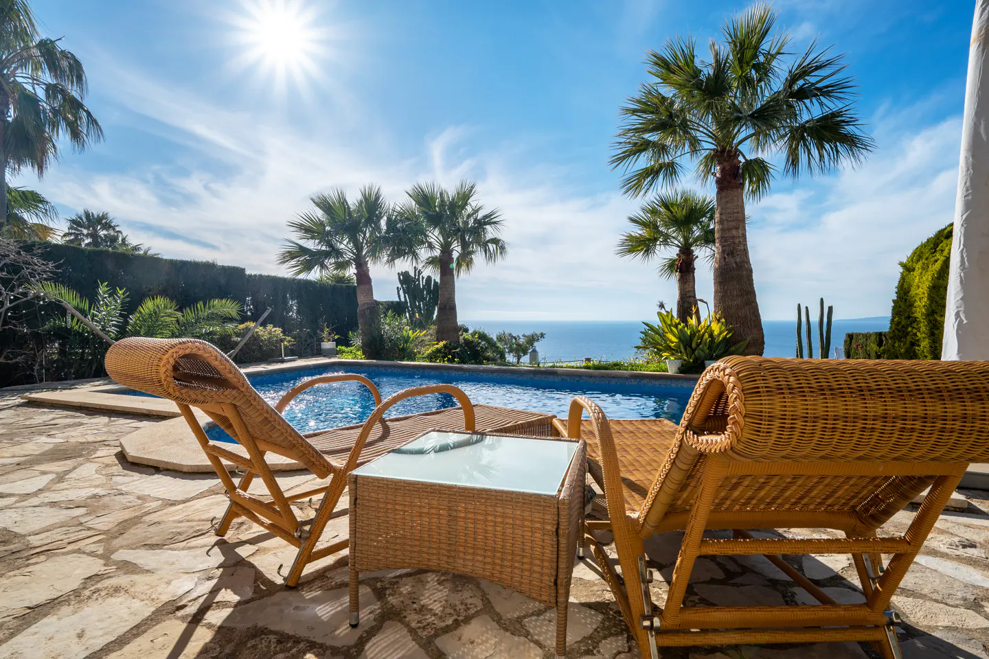 Outdoor patio with pool, ocean view, and palm trees. Two wicker lounge chairs and a table sit on a stone patio under a sunny blue sky.