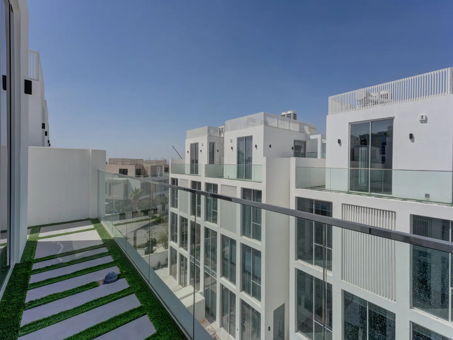 View from a balcony with glass railings, overlooking modern white buildings under a clear blue sky. The balcony has gray tiles with artificial grass.
