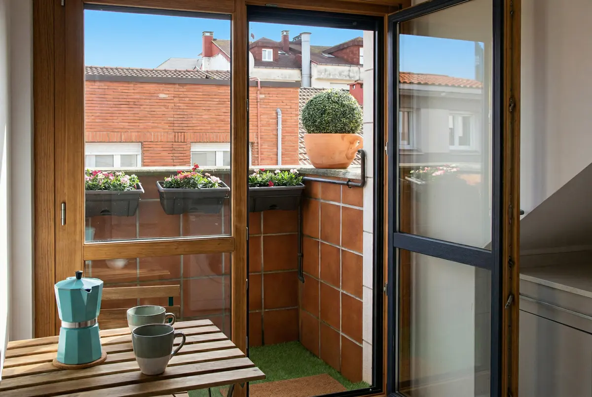 A cozy balcony with a wooden table set, a blue moka pot, and two mugs. The balcony has terracotta tiles and flower boxes with colorful flowers.