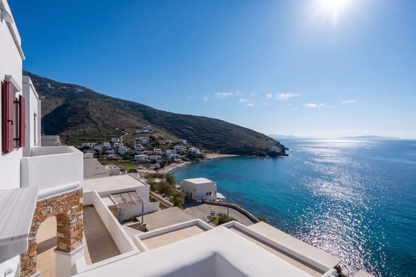 View from a white building overlooking a turquoise bay, a sandy beach, and a hillside village under a clear blue sky.