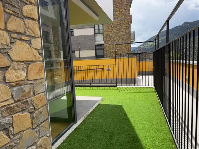 A balcony with artificial green grass, black metal railings, and a stone wall. A yellow building is in the background.