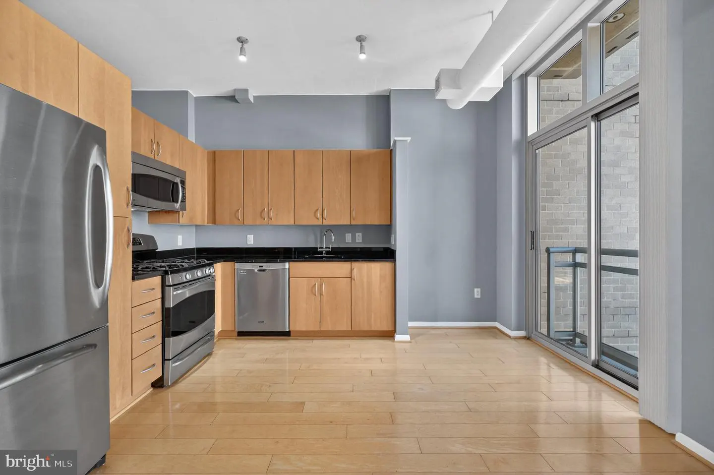 A modern kitchen with stainless steel appliances, light wood cabinets, black countertops, and a sliding glass door to a balcony.