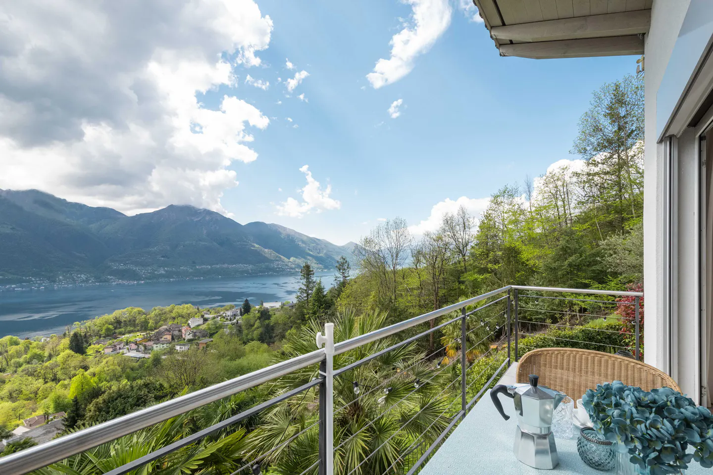 Balcony view of mountains and lake. Table with coffee pot and blue flowers. Railing in foreground.