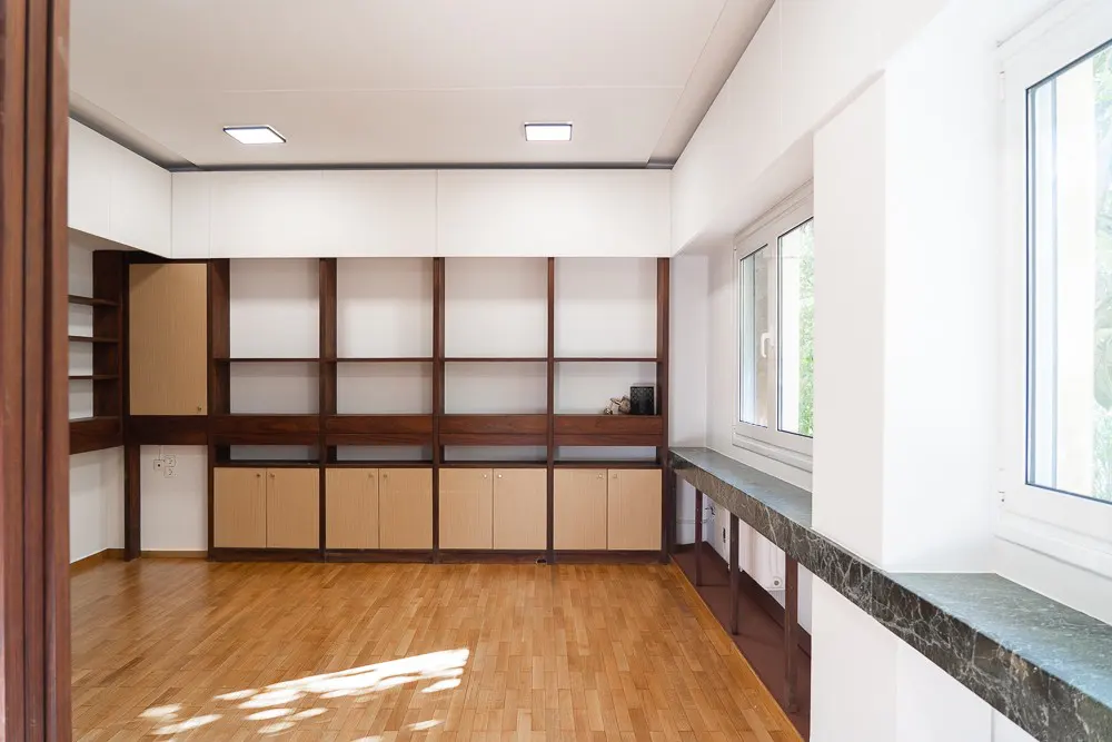 Bright room with wood floors, white walls, and built-in shelving. Dark wood frames open shelves and tan cabinets. Windows with marble sills let in light.