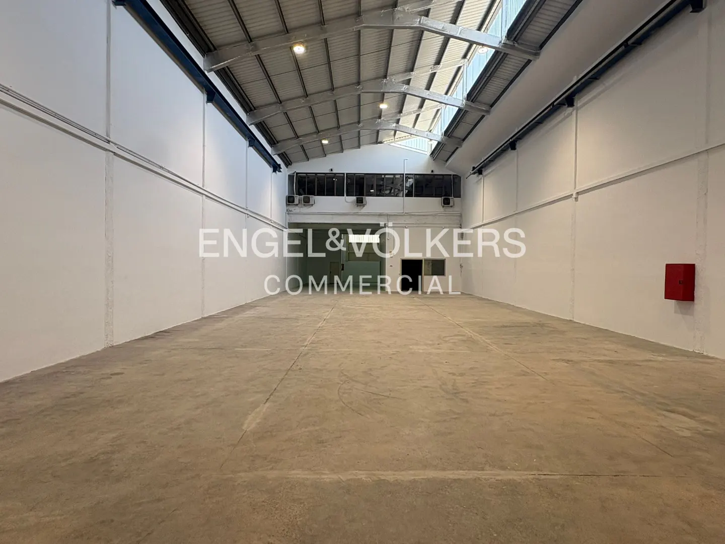 Empty, spacious industrial warehouse with high ceilings, white walls, concrete floors, and a mezzanine office area.