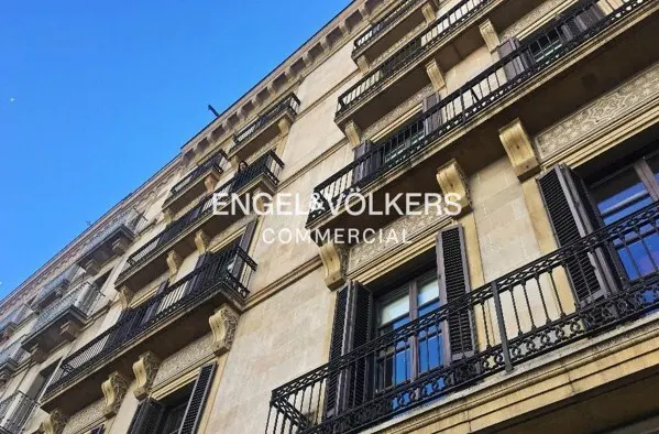 Exterior view of a beige building with black iron balconies and shutters under a blue sky. "Engel & Völkers Commercial" is printed on the building.