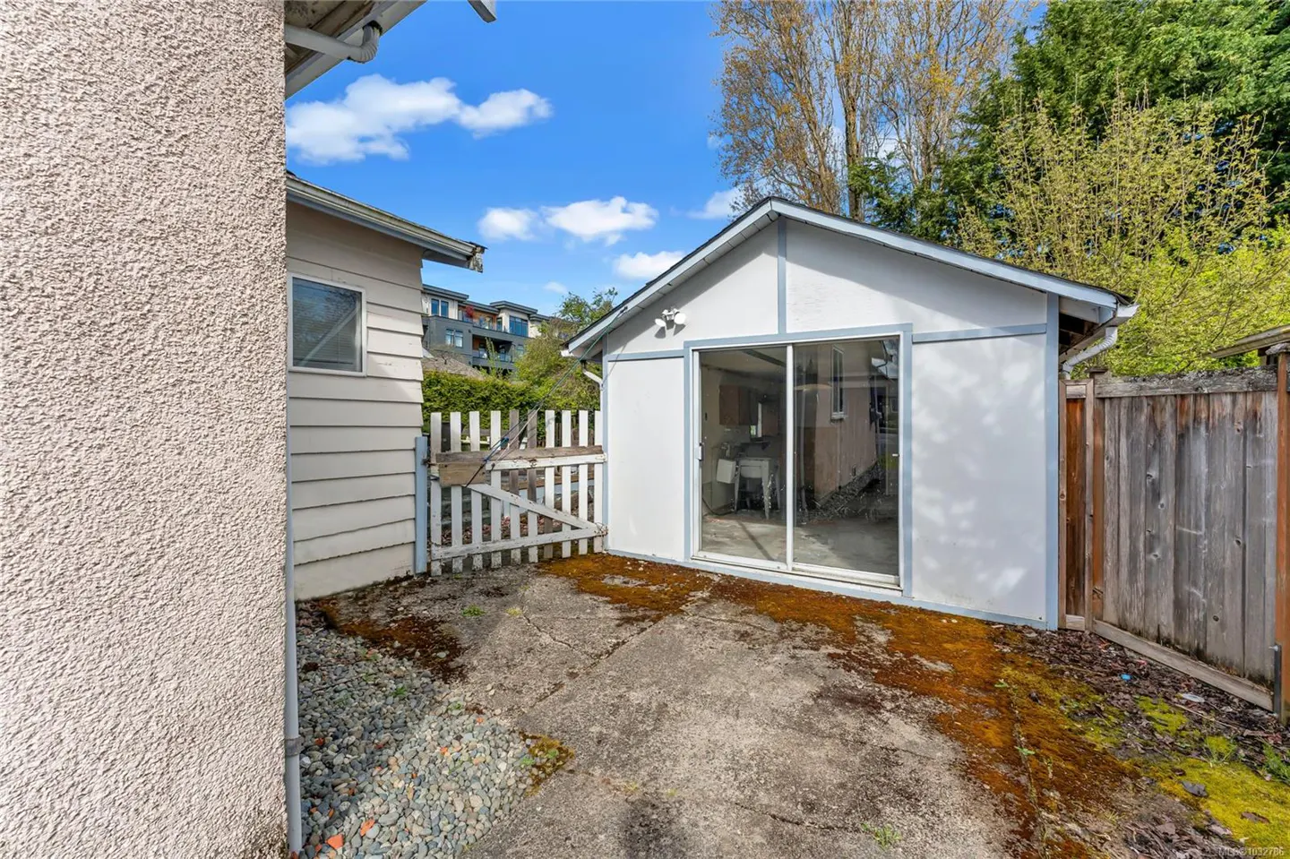Exterior view of a white shed with a sliding glass door, a white picket fence, and a concrete patio with moss.