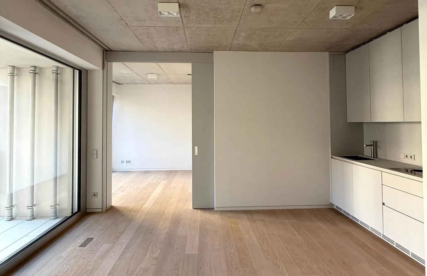 Modern apartment interior with light wood floors, white minimalist kitchen cabinets, and a concrete ceiling.