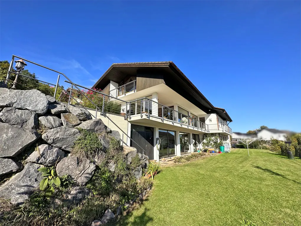 Exterior view of a two-story house with a brown roof, white walls, and balconies, set on a green lawn under a clear blue sky. A rock wall and stairs lead to the house.