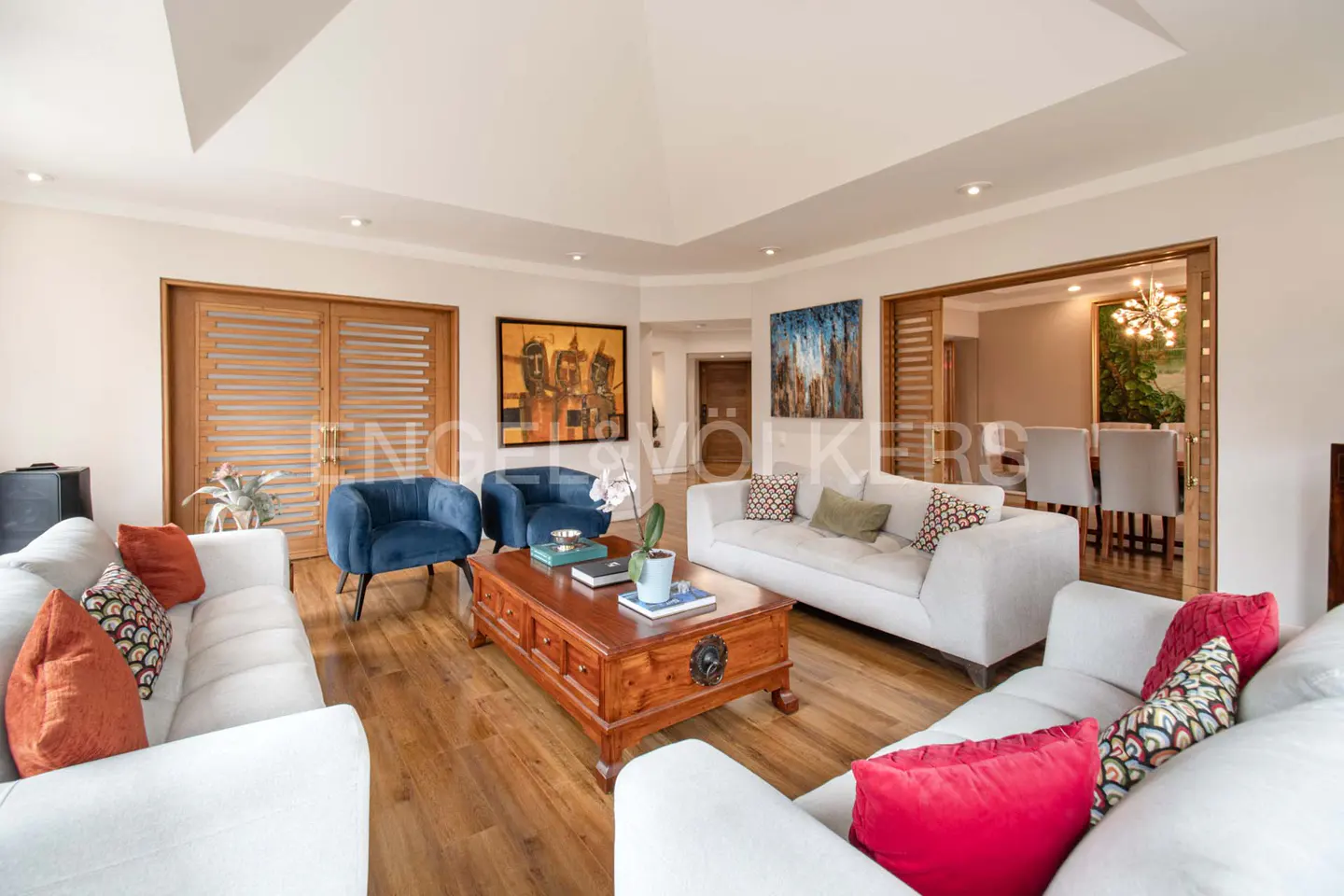 Bright living room with white sofas, blue chairs, and a wooden coffee table on a wood floor. Artwork hangs on the walls.
