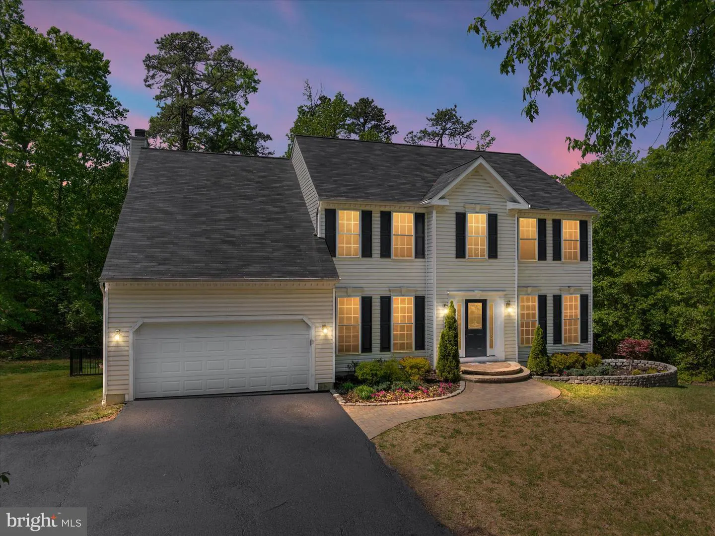 Two-story house with white siding, black shutters, and a gray roof at dusk. A driveway leads to an attached garage.