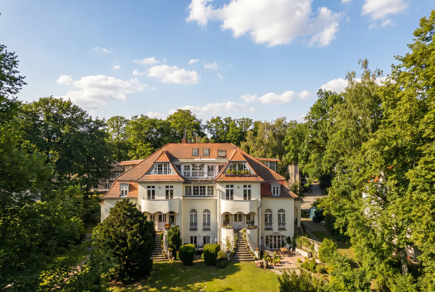 A large, cream-colored house with a red tile roof is surrounded by green trees under a blue sky with white clouds.