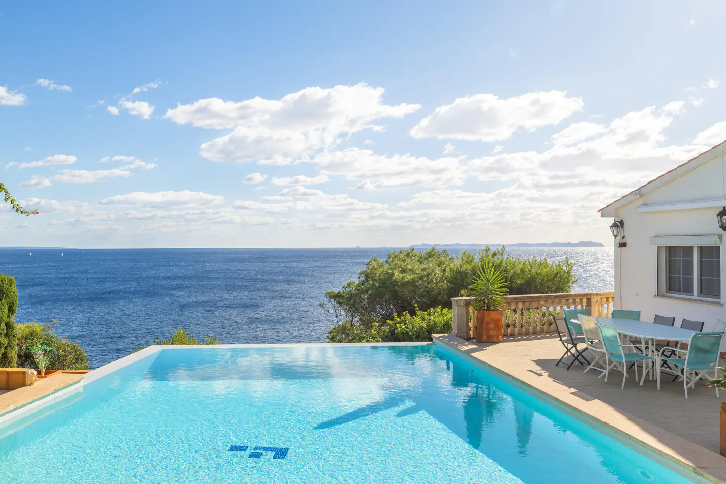 Infinity pool overlooking the ocean, next to a white house with a patio and outdoor dining set.