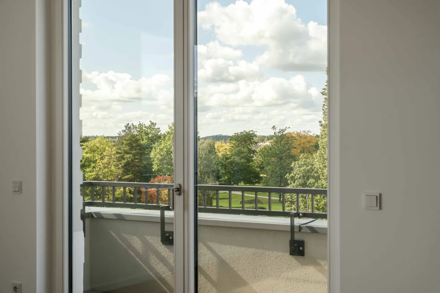 View from a bright room through a glass door onto a balcony overlooking a lush green park with trees under a cloudy sky.
