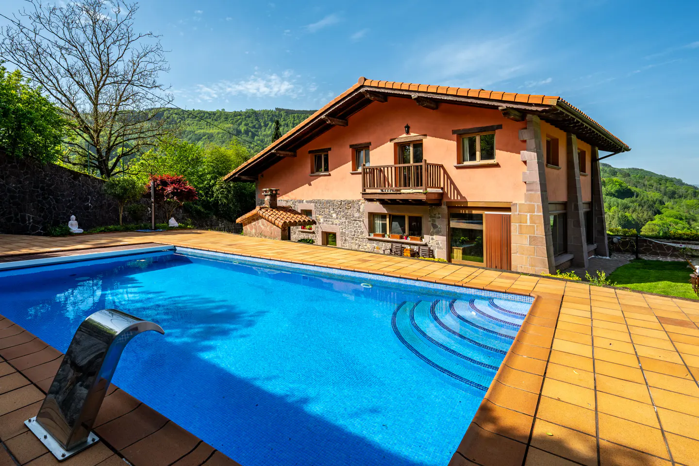 A two-story house with a pool. The house is peach-colored with a brown roof and a wooden balcony. The pool is blue with mosaic steps and a metal waterfall feature.