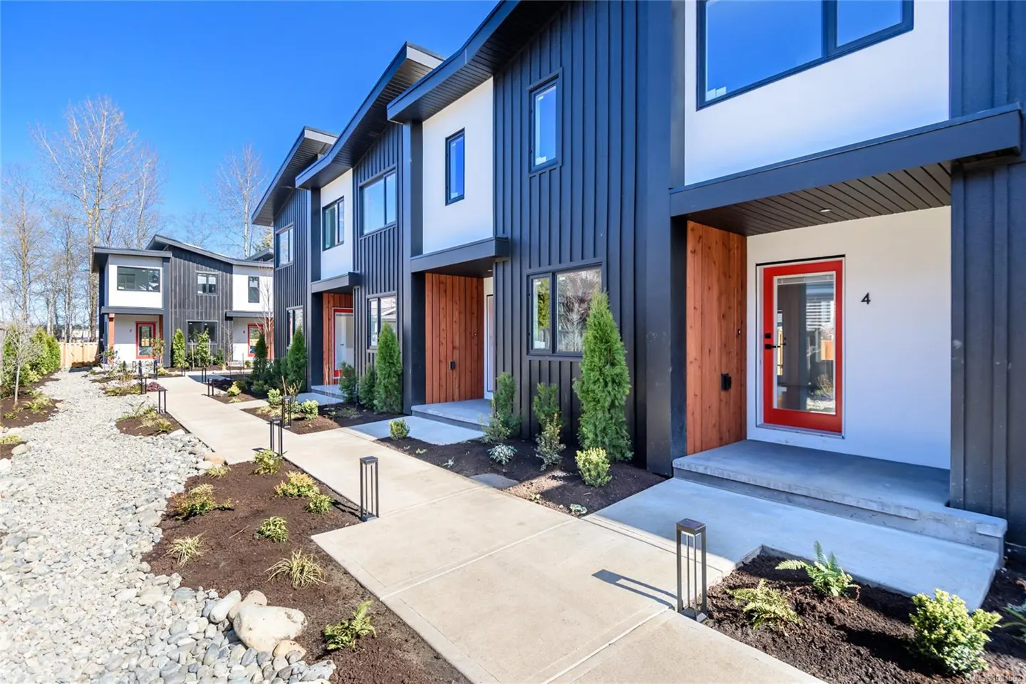 Row of modern townhouses with black siding, white accents, and red doors. A concrete walkway leads to each unit.