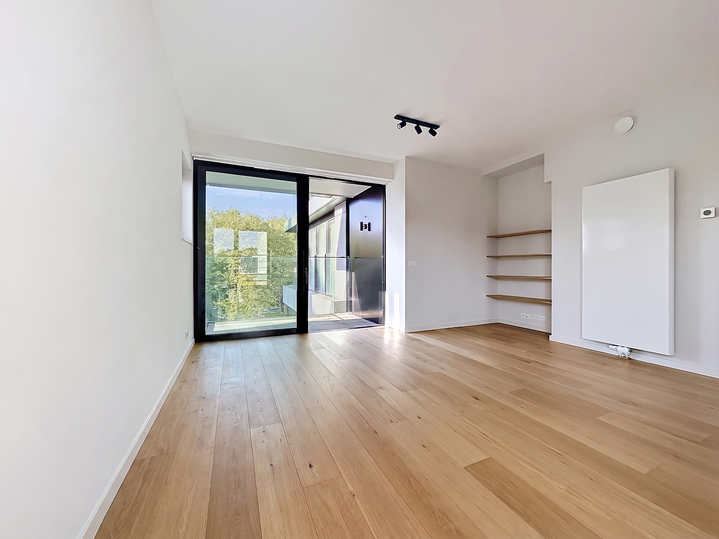 Bright, empty room with wood floors, white walls, and a black-framed sliding glass door to a balcony. Shelves and a radiator are on the right wall.