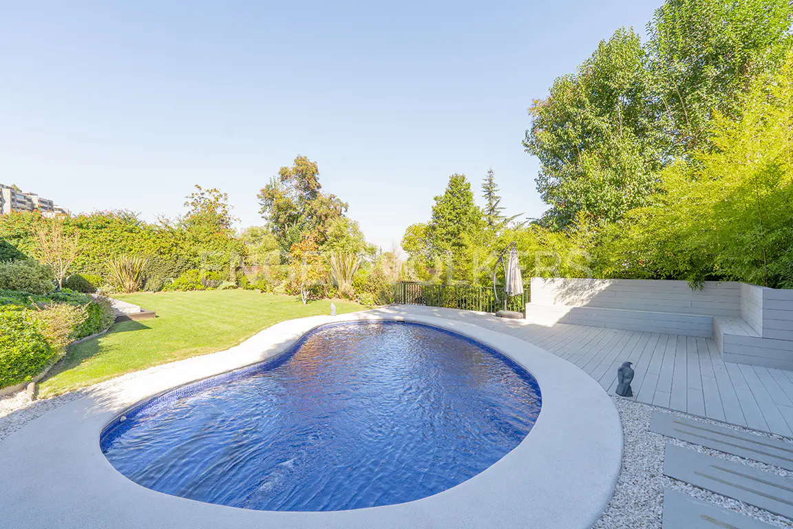 A kidney-shaped blue pool with a white border is surrounded by a green lawn, trees, and a gray deck.