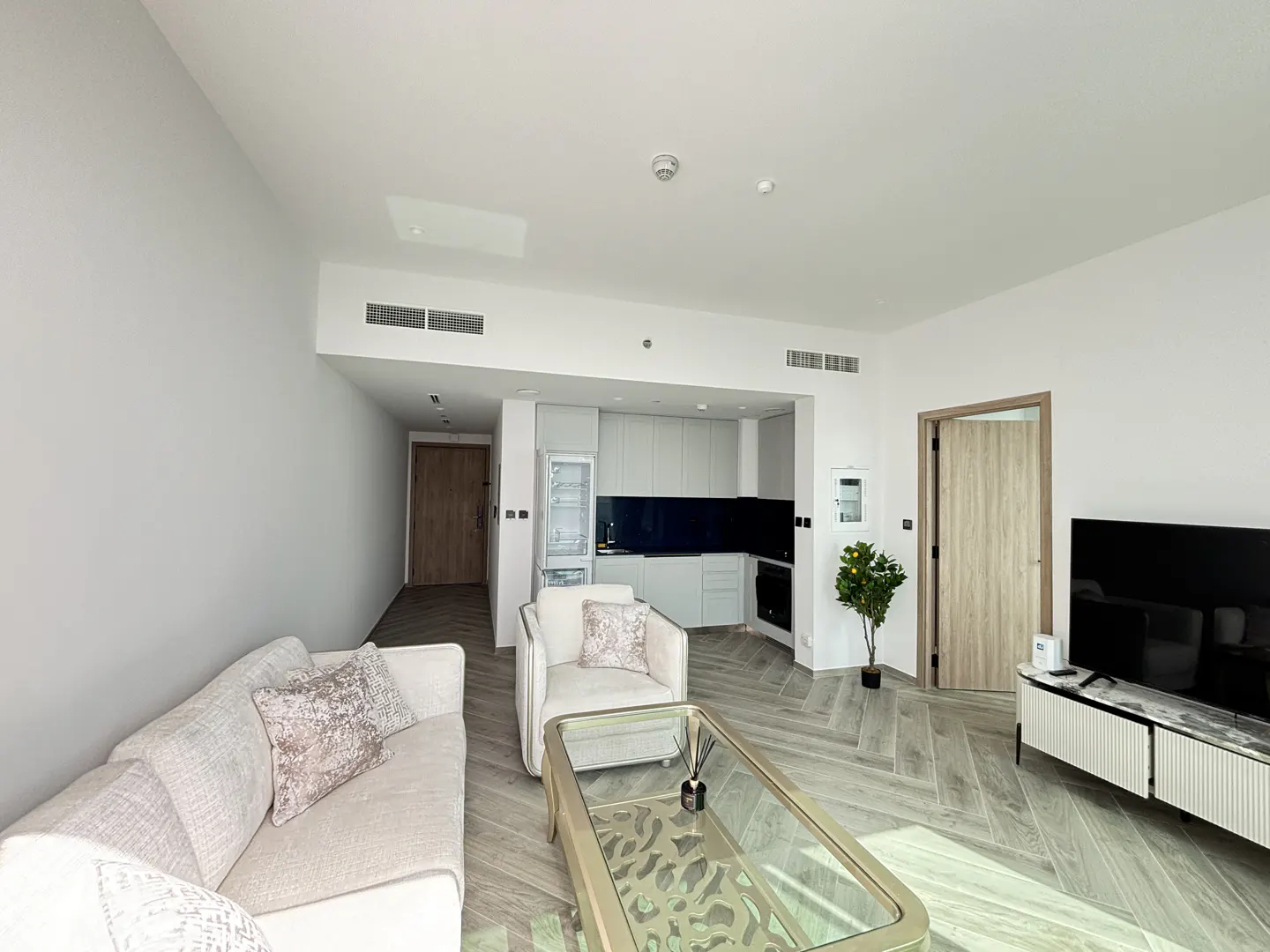 Bright, modern apartment living room with white sofa, chair, glass table, and kitchen in the background. Herringbone wood floors.