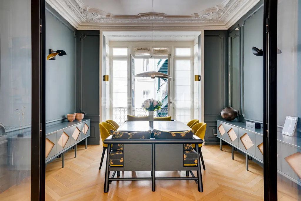 Dining room with herringbone wood floors, gray walls, and white trim. Table with yellow chairs and floral bench. Large window with natural light.