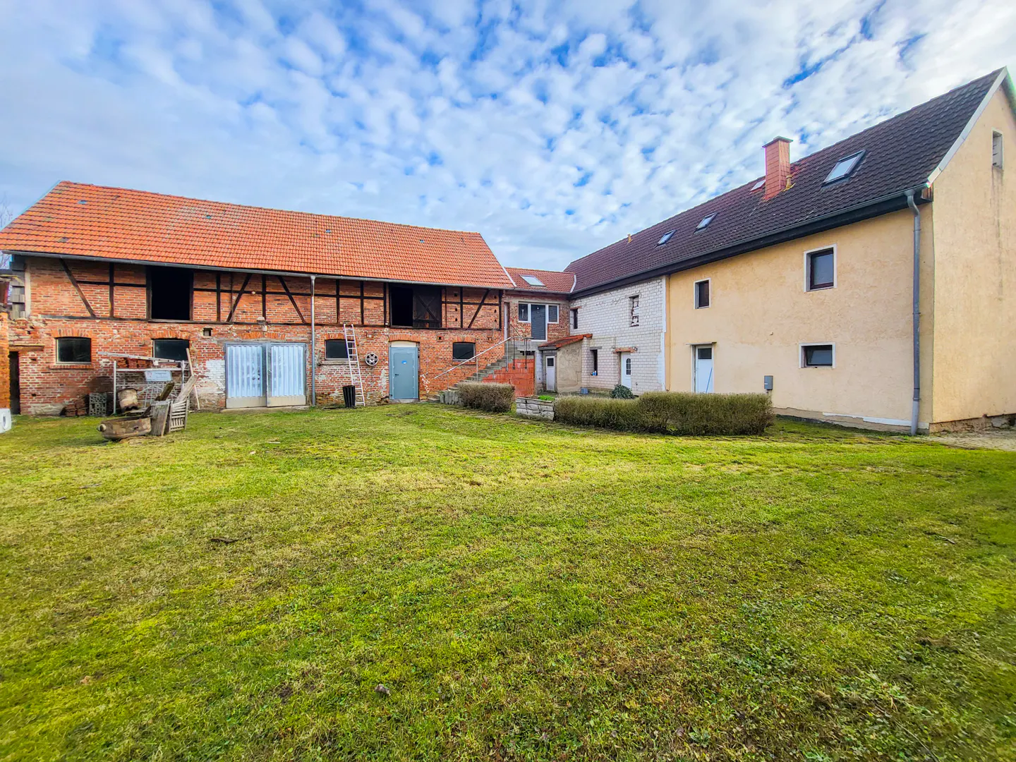 Exterior view of a large, multi-building farm complex with a green lawn and cloudy sky. The buildings are brick and stucco with red tile roofs.