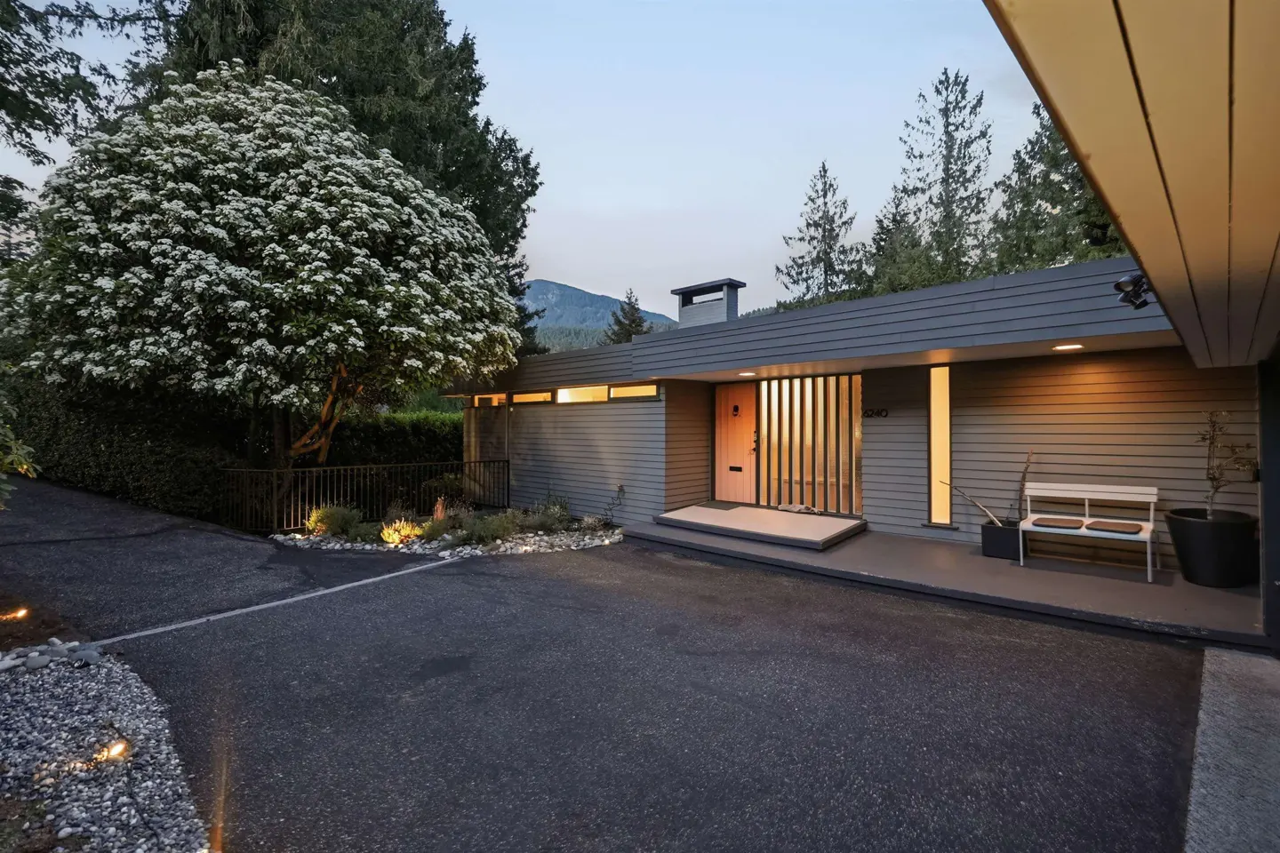 Exterior of a gray, modern home with a white bench on the porch and a tree with white flowers.