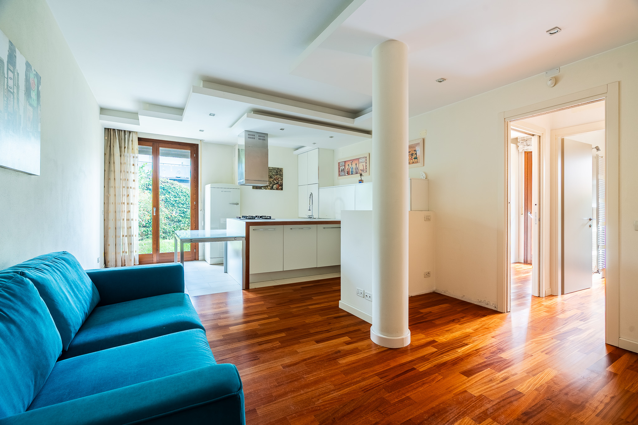 Bright open-plan living area with a blue sofa, hardwood floors, a modern white kitchen, and a large central support pillar. [AI-generated text]