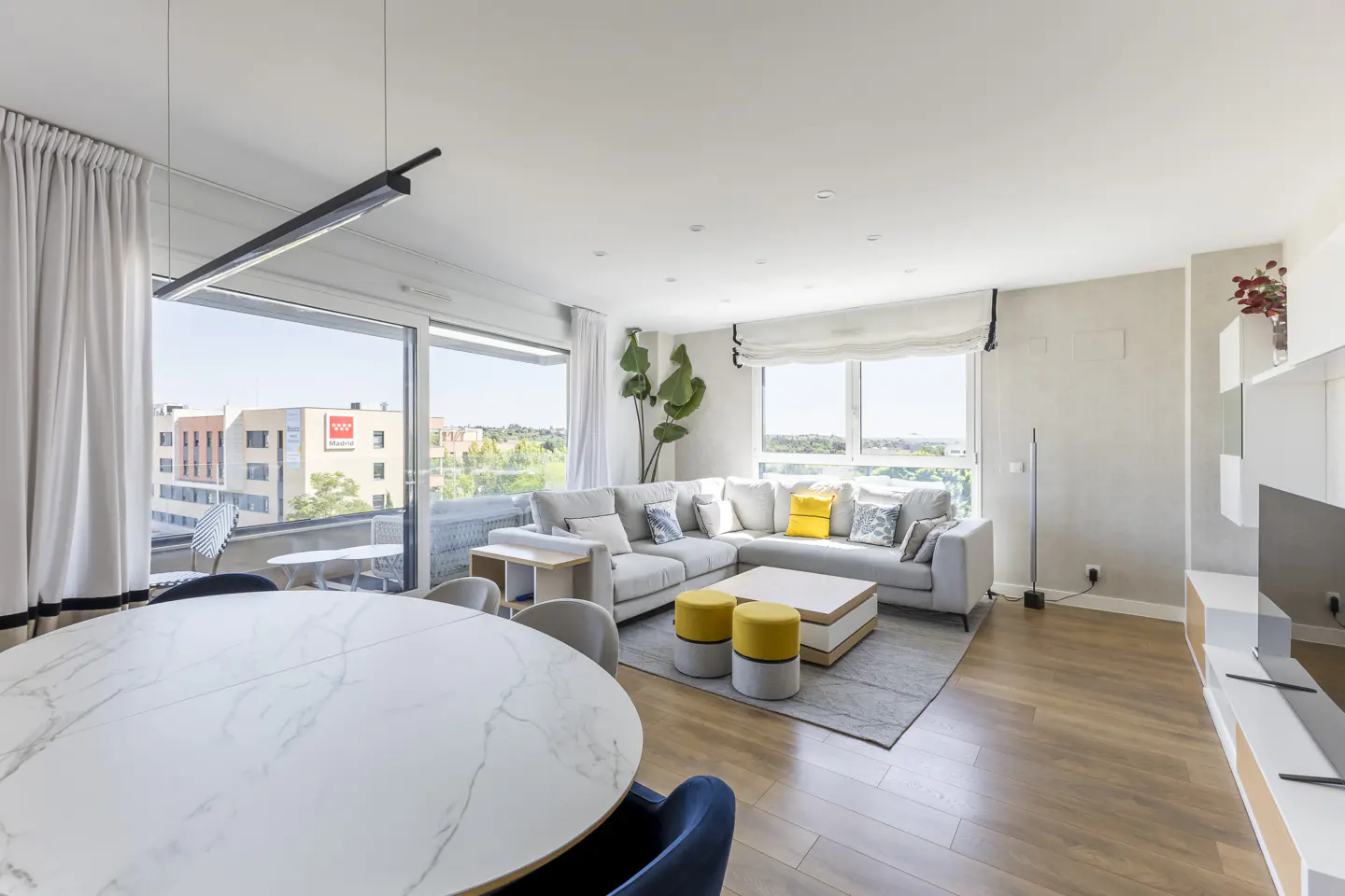 Bright, modern living room with a white marble table, gray sectional sofa with yellow accents, and a large window with city views.