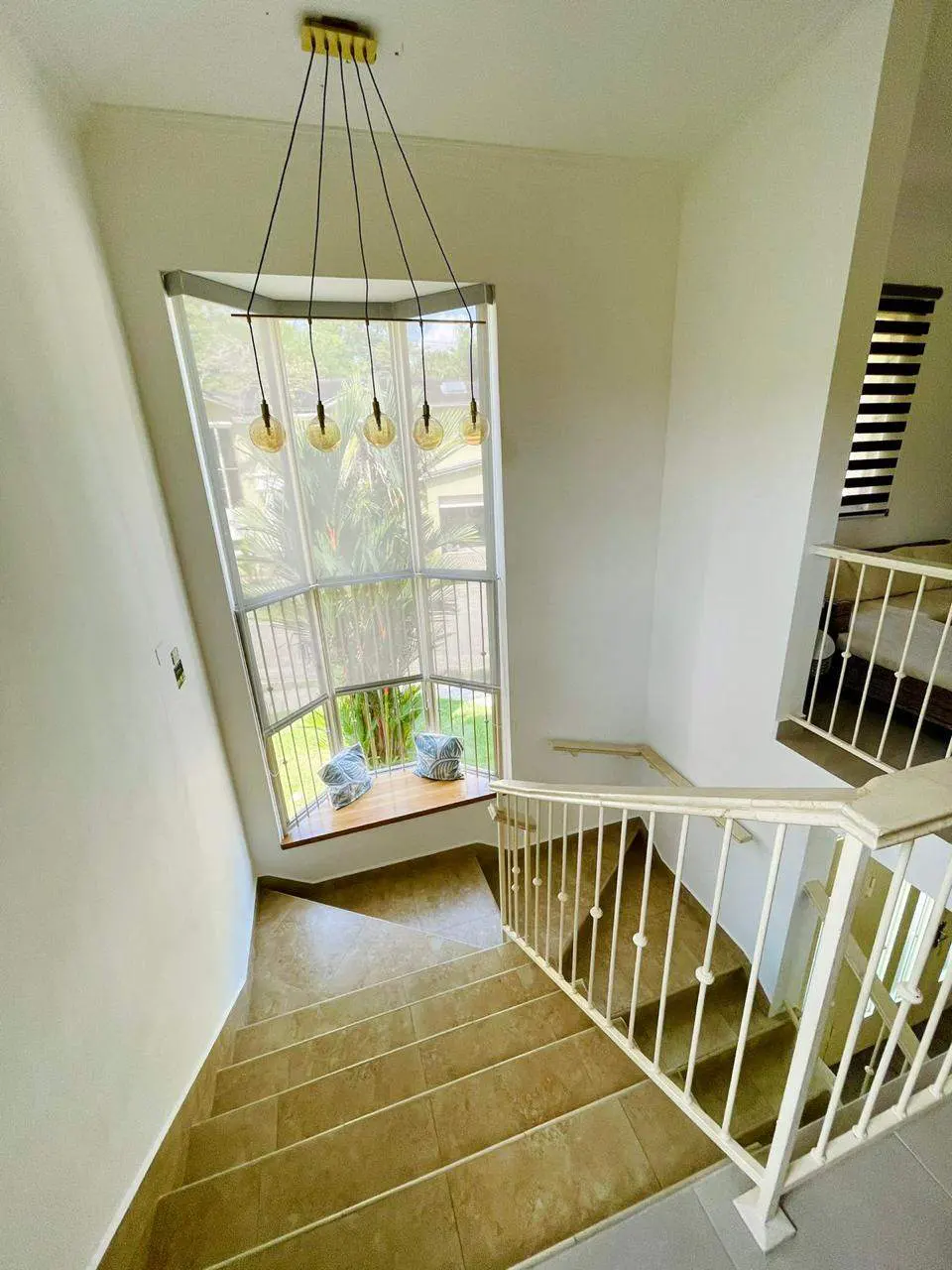 Interior view of a staircase with beige tile steps and white railing, leading down from a window with a view of greenery. A modern chandelier hangs above.